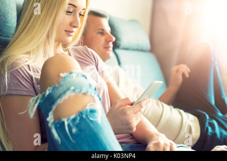 Young stylish couple sitting on a sofa relaxing at their modern home. Woman holding mobile phone, browsing the web, man watching television Stock Photo