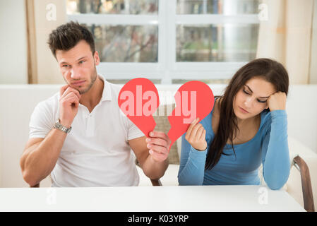 Portrait Of A Sad Couple Sitting On Chair Holding Red Broken Heart Stock Photo