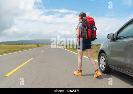 elegant leisurely female backpacker standing on asphalt road watching distant mountain scenery and parked car in roadside at summer vacation travel. Stock Photo