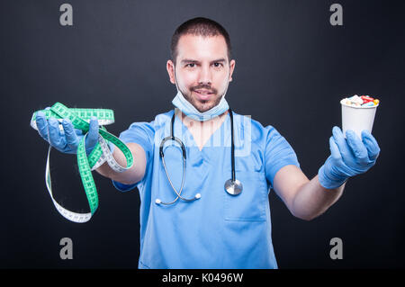 Plastic surgeon wearing scrubs holding in balance measuring tape and pills smiling on black background Stock Photo
