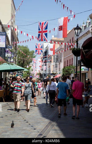 People strolling along Main Street in the UK dependency of Gibraltar with the Union Jack and the flag of Gibraltar in the background Stock Photo