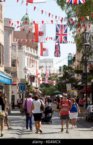 People strolling along Main Street in the UK dependency of Gibraltar with the Union Jack and the flag of Gibraltar in the background Stock Photo