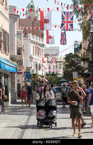 People strolling along Main Street in the UK dependency of Gibraltar with the Union Jack and the flag of Gibraltar in the background Stock Photo