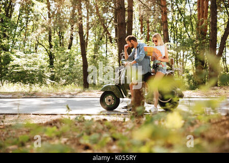 Image of young tired loving couple holding map outdoors sitting on scooter. Looking aside. Stock Photo