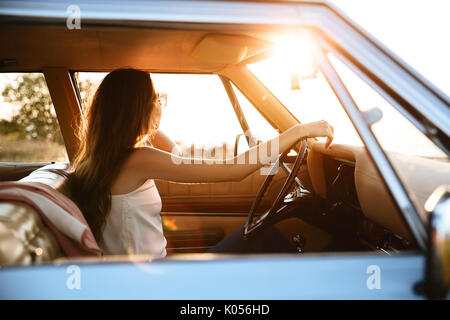 Side view of a young woman sitting inside a retro car and holding hands on a steering wheel Stock Photo