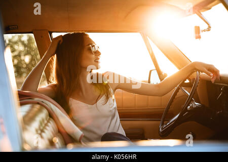 Side view of a young smiling woman in sunglasses sitting inside a retro car and holding hands on a steering wheel Stock Photo