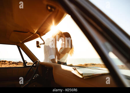 Young happy couple leaning on a car while standing and hugging outdoors in sunlight Stock Photo