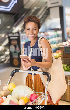 Portrait smiling young woman using cell phone, grocery shopping in market Stock Photo