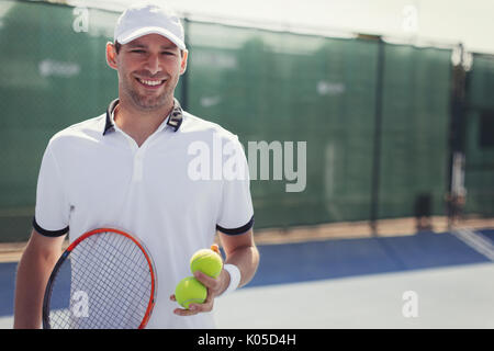 Portrait confident, smiling young male tennis player holding tennis racket and tennis balls on sunny tennis court Stock Photo