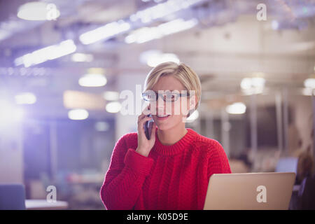 Businesswoman talking on cell phone at laptop in dark office, working late Stock Photo