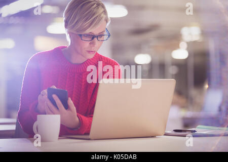 Serious businesswoman working late, using cell phone at laptop in office Stock Photo