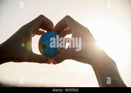 closeup of a young caucasian man with a world globe in his hands forming a heart against the sky and a ray of sun showing up on the right Stock Photo
