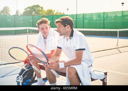 Young male tennis players resting with tennis rackets on sunny tennis court Stock Photo