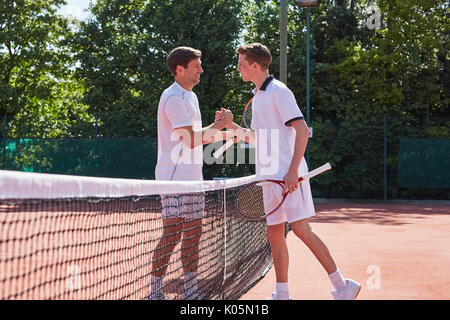Young male tennis players shaking hands in sportsmanship over net on sunny clay tennis court Stock Photo