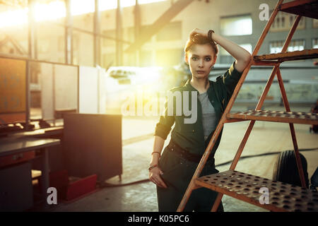 Portrait of modern young woman in green uniform working in engineering standing by jet plane near the stairs looking at camera. Stock Photo