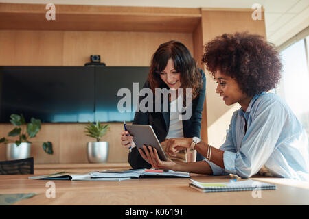 African businesswoman showing something on digital tablet to her female colleague while sitting at her desk. Professional employees discussing ideas o Stock Photo