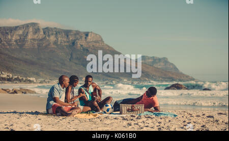African man playing guitar for his friends along the sea shore. Group of people relaxing on the sandy beach. Stock Photo