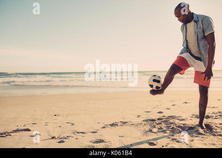 Afro american man playing with football along the beach. Black male balancing a soccer ball on his feet at the sea shore. Stock Photo