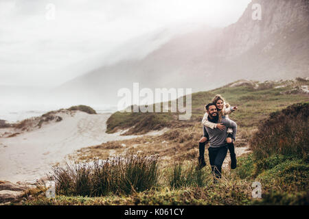 Outdoor shot of man giving woman piggyback on winter beach. Man carrying woman oh his back pointing away.