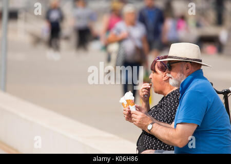 Mature couple retired enjoying a cooling ice cream and cone sat down on the promende on a summer day at the coastal resort seaside town of Colwyn Bay in North Wales, UK Stock Photo