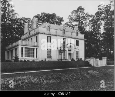 Exterior of a grand home, featuring chimneys, dormer and standard windows, an enclosed porch, a second story balcony, and two small sets of stairs leading to a covered entryway, situated in a landscaped lawn with a pathway, in a secluded area on a quiet street in Baltimore, Maryland, 1910. This image is from a series documenting the construction and sale of homes in the Roland Park/Guilford neighborhood of Baltimore, a streetcar suburb and one of the first planned communities in the United States. Stock Photo