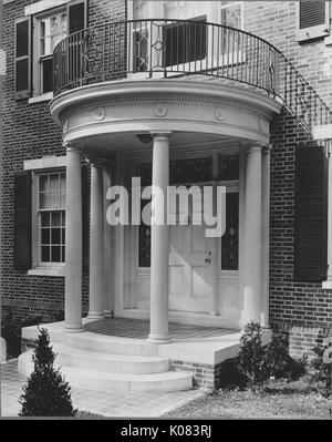 Partial view of a brick home that features windows with shutters, a second-story front balcony held up by classical columns that form the entryway to the home, a set of three steps that lead to the front door, and sparse landscaping including shrubbery, in Baltimore, Maryland, 1910. This image is from a series documenting the construction and sale of homes in the Roland Park/Guilford neighborhood of Baltimore, a streetcar suburb and one of the first planned communities in the United States.