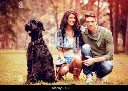 Beautiful lovely couple with a dog, a black giant schnauzer, enjoying in the park in autumn colors. Looking at camera. Stock Photo