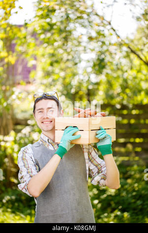 Photo of brunette in gloves holding box of carrots on shoulder Stock Photo
