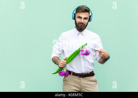 Steep young man with blue headset and holding arms penny skateboard. Light green background. Studio shot Stock Photo