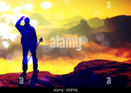 Film grain effect. Silhouette of tourist with poles in hand. Hiker stand on rocky view point above misty valley. Sunny spring daybreak in rocky mounta Stock Photo