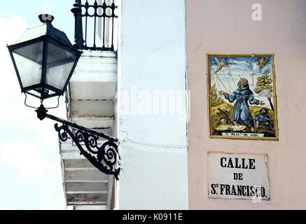 A religious artwork of ceramic tiles marks Calle de San Francisco, a street named for Saint Francis of Assisi in historic Old San Juan in Puerto Rico (PR), an unincorporated territory of the United States in the Caribbean Sea. A vintage wrought iron street light illuminates the street name and illustrated sign at night. Stock Photo