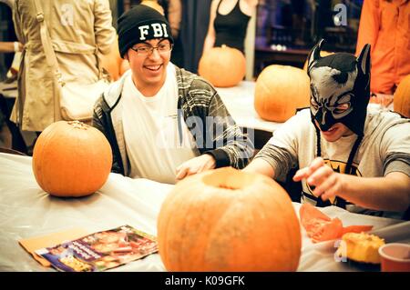 Two college students are sitting at a table each with a pumpkin, one is carving their pumpkin and the other pumpkin has not been touched yet, the students are smiling and are also in costume, one is wearing an FBI hat and the other is wearing a Batman hat and mask, Halloween at Johns Hopkins University's George Peabody Library, 2015. Courtesy Eric Chen. Stock Photo