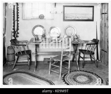 Landscape shot of a wooden table with three different types of wooden chairs; three plate a cup sets lay on the table and propped up against the wall; two different-seized patterned rugs on the ground, a landscape photo hanging on the ceiling next to a large window; Roland Park/Guilford, Baltimore, Maryland, 1910. This image is from a series documenting the construction and sale of homes in the Roland Park/Guilford neighborhood of Baltimore, a streetcar suburb and one of the first planned communities in the United States. The neighborhood was segregated, and is considered an early example of t Stock Photo