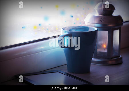 Winter decoration with lantern, hot tea cup,e-book reader and lights on window . Stock Photo