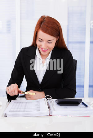 Smiling young businesswoman examining documents with magnifying glass at office desk Stock Photo
