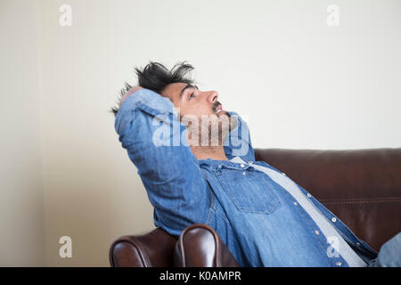 Side view of a tired or unhappy Asian man sitting on sofa holding his head. Stock Photo