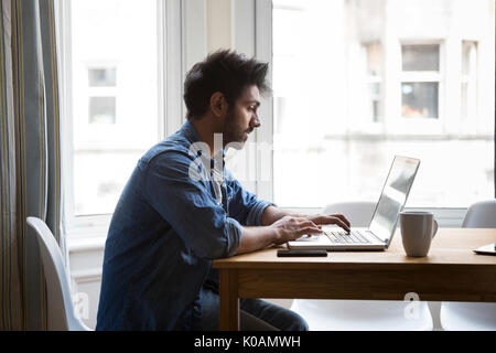 Portrait of an Indian man sitting at a table at home working on a laptop computer. Side View. Stock Photo