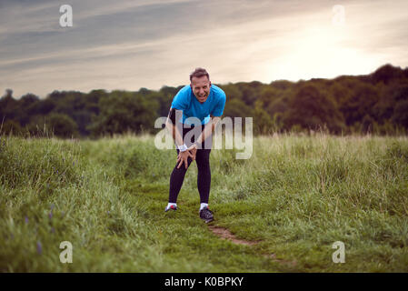 An exercising man in fit wear while running outdoors, stop to clutch an injured knee. Stock Photo