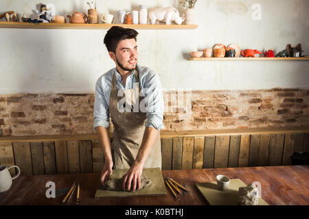 potter, workshop, ceramics art concept - standing young smiling brunette man with hands knead fireclay, male dressed in apron, a ceramist with raw materials on wooden table with sculpting tools set. Stock Photo