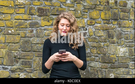 A pretty young woman using her thumbs to enter text and send a message from her mobile phone, while standing in front of a brick wall. Stock Photo