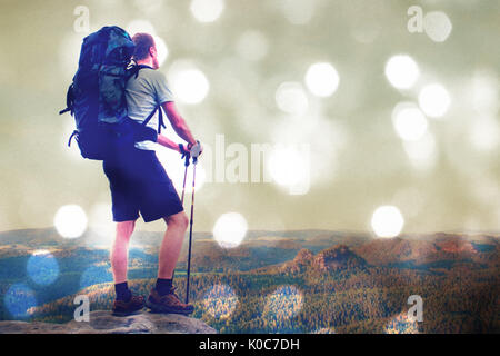 Film grain effect. Tall tourist with poles in hand. Sunny evening in rocky mountains. Hiker with big backpack stand on rocky view point above misty va Stock Photo