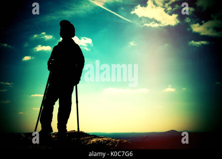 Film grain effect. Adult hiker with poles in hand. Hiker take a rest on rocky view point above valley. Sunny spring daybreak in rocky mountains. Stock Photo