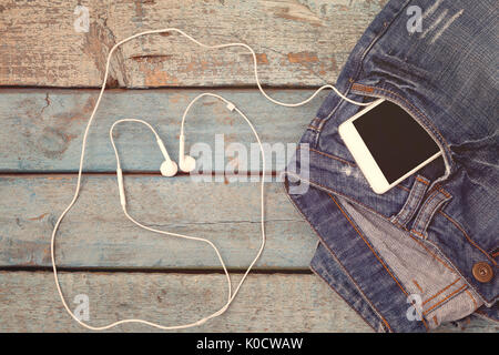 Touch smartphone-phone with headset-headset lined in the shape of a heart in a jeans pocket on a blue wooden background. Stock Photo