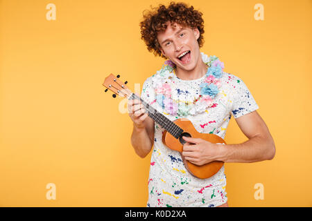 Attractive young man in summer clothes playing ukulele and singing isolated over yellow background Stock Photo