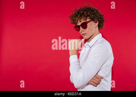 Portrait of a young thoughtful stylish man in sunglasses standing over red background Stock Photo