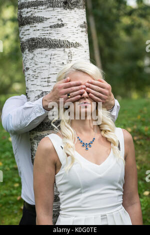 Couple in love. Man covered eyes of smiling blonde woman by his hands in park. Young people resting in nature, girl leaned against a birch tree Stock Photo