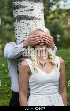 Couple in love. Man covered eyes of smiling blonde woman by his hands in park. Young people resting in nature, girl leaned against a birch tree Stock Photo