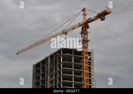 Upper part of a constructed tall building with a tower crane jib over the roof against dramatic cloudy sky. Stock Photo