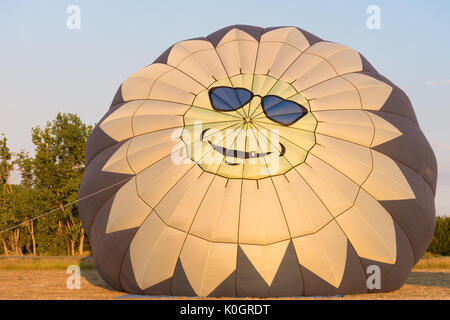A hot air balloon with a happy sun face with blue sunglasses on the top, The balloon is being inflated at sunrise. Stock Photo