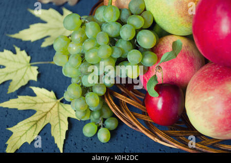 Harvest of ripe fruits - apples, grapes, plums, pears in a large wooden plate on a black background, decorated with autumn maple leaves. Harvest conce Stock Photo